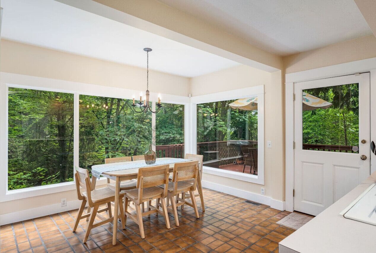 Un espace repas comprenant une table et des chaises, de grandes fenêtres donnant sur un paysage verdoyant et une porte offrant une vue sur les arbres.