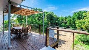 Een houten terras met een tafel en stoelen, een overdekt gedeelte met dak, en uitzicht op weelderig groen.