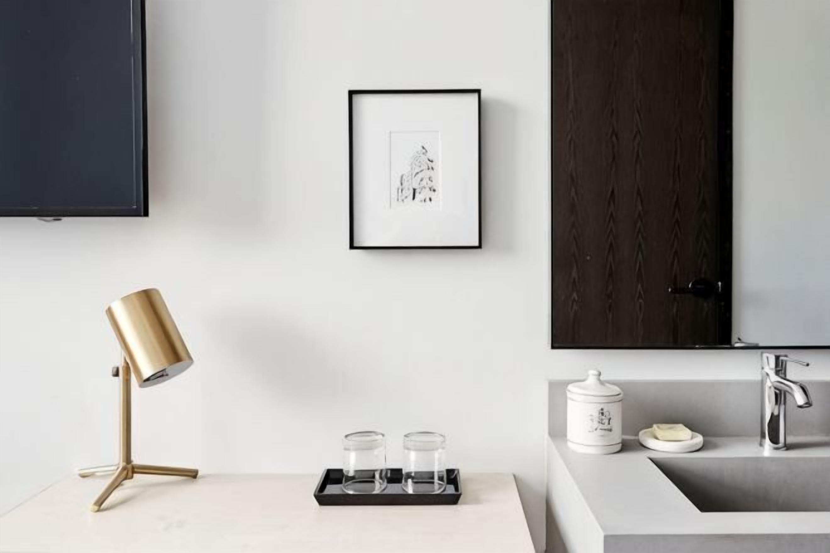 Une salle de bain moderne avec un comptoir blanc, une lampe dorée, un tableau encadré, un lavabo avec un porte-savon et une porte en bois foncé.