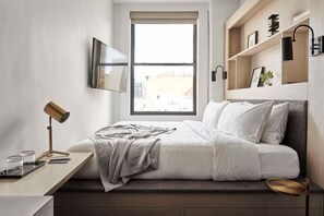 Een slaapkamer met een bed, een bureau, een stoel, een raam en een plank met decoratieve objecten.