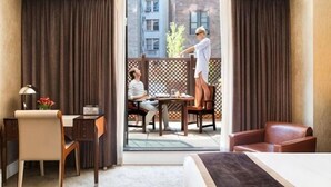 A hotel room with a bed, a desk with a lamp, a chair, and a view of a balcony with two people.