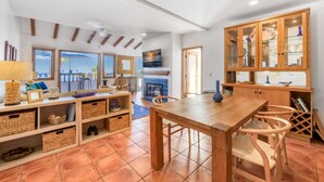 Un vaste espace de vie comprenant une table et des chaises en bois pour la salle à manger, une cheminée et une vue sur la mer grâce à de grandes fenêtres.