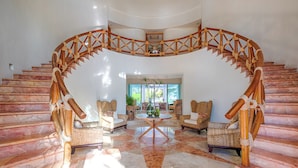A grand staircase with a wooden balustrade leading to a balcony, surrounded by wicker furniture and a marble floor.