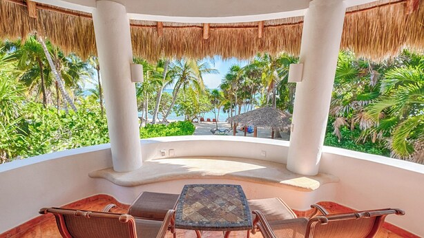 A covered outdoor seating area with a table and chairs, overlooking a beach with palm trees and a thatched-roof structure.
