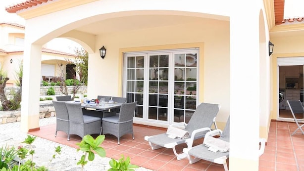 A covered patio with a table and chairs, a sliding glass door, and a tiled floor. - Casa América - Praia Del Rey (Casal da Lagoa Seca)