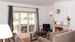 A living room with a sofa, a coffee table, a TV, and a fireplace. There is a sliding glass door leading to an outdoor area with furniture. - Casa América - Praia Del Rey (Casal da Lagoa Seca)