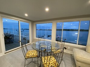 Un comedor con mesa y sillas de vidrio, ventanas amplias con vista al agua, y una terraza con mobiliario exterior.