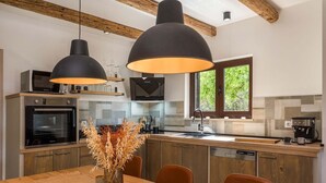 A modern kitchen with wooden beams, two pendant lights, a built-in oven, a coffee machine, and a dining area with a table and chairs.