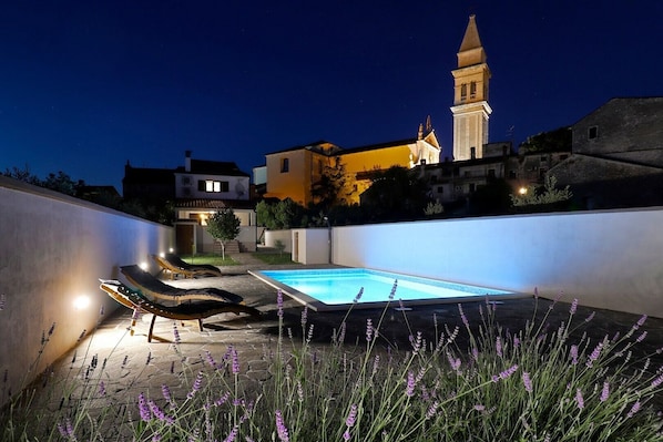 Vista nocturna de una piscina con sillas de descanso y plantas de lavanda, rodeada por un muro bajo y una torre de iglesia al fondo.