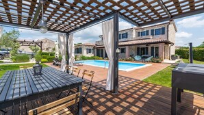 A covered outdoor area with a pool, wooden deck, and a stone building in the background. - Rustic Villa Noce with pool (Sveti Ivan)