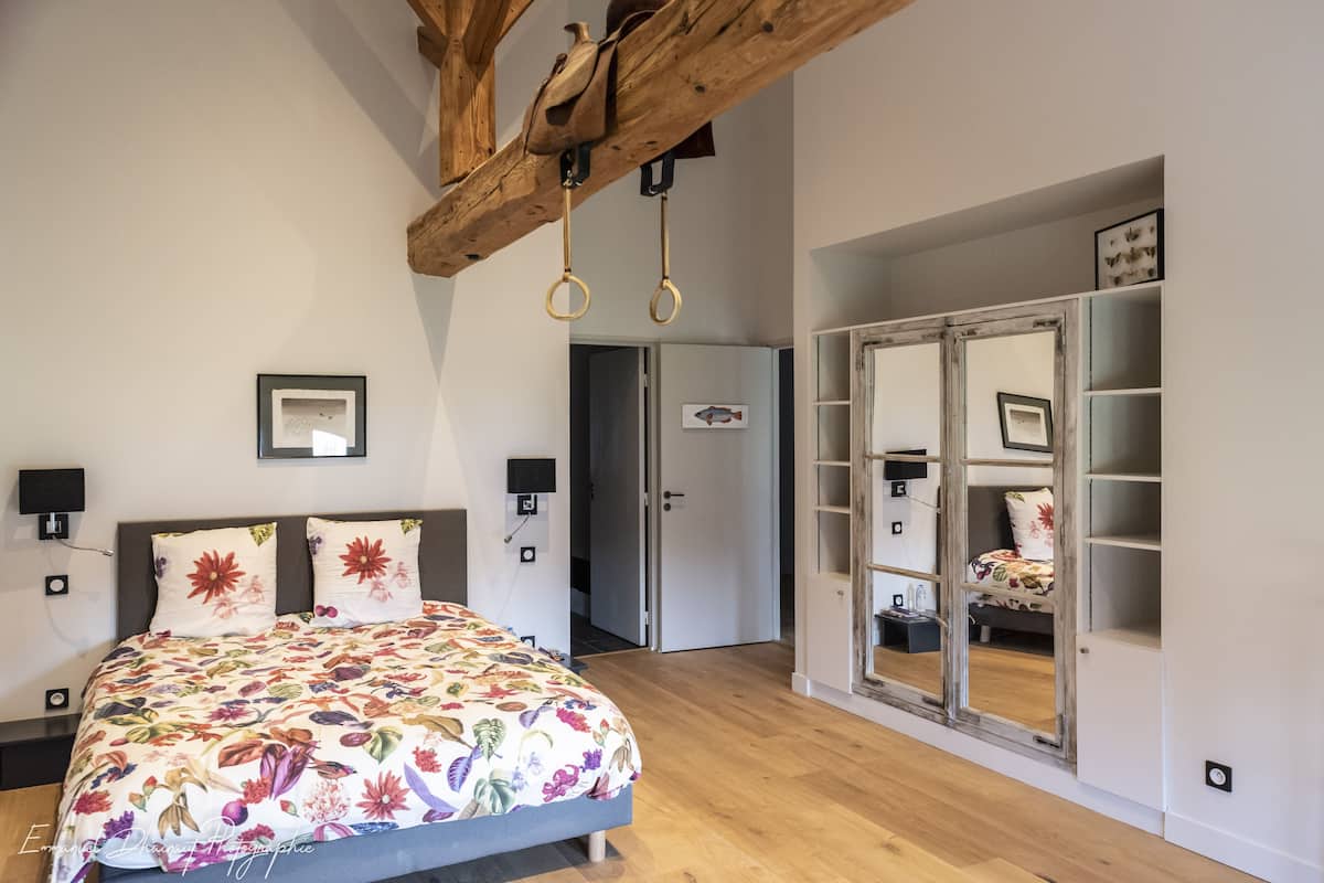 A bedroom with a floral bedspread, a wooden ceiling, a wardrobe with a mirror, and a door leading to another room.