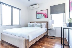 Een slaapkamer met een houten bed, witte beddengoed, een nachtkastje met een lamp en een potplant.
