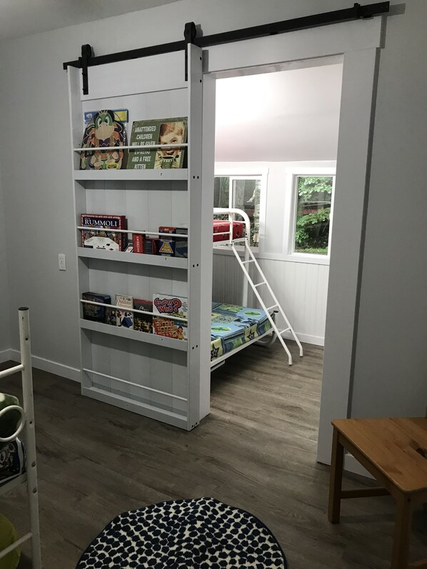 Una habitación con una estantería blanca llena de juegos de mesa, una litera con escalera y una mesa de madera.