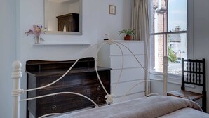 Ein Schlafzimmer mit einem hölzernen Bettgestell, einem Schminktisch, einem Spiegel, einem Fenster mit Vorhängen und Blick ins Freie.