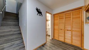 Un couloir avec un escalier en bois, une décoration murale représentant un cerf et une armoire en bois dotée de portes à persiennes.