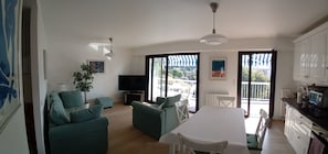 A spacious living area with a sofa, a dining table, and a TV. There are large windows and a view of the outside.