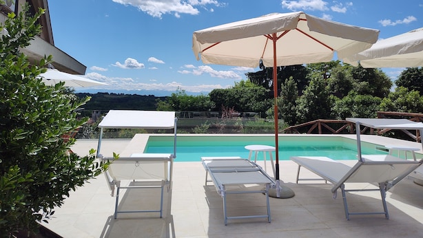A poolside area with lounge chairs and umbrellas, a clear blue sky, and a view of a pool and greenery in the background.