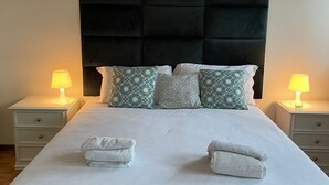 A neatly made bed with white linens and decorative pillows, flanked by two bedside tables with lamps, in a room with a dark headboard and wooden flooring.