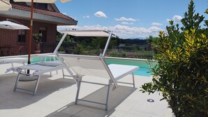 A poolside area with lounge chairs and umbrellas, a brick building in the background, and a clear sky.