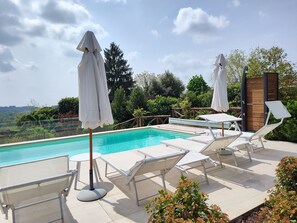 A poolside area with lounge chairs and umbrellas, surrounded by greenery and a wooden fence.