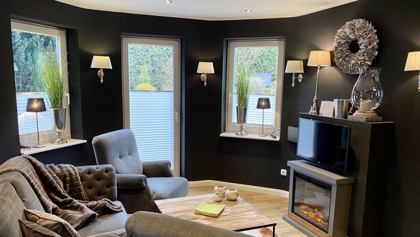 A modern living room with a grey sofa, a television, and a fireplace. There are two windows with plants and lamps, and a dark wall with a wreath. - country-apartment XL for 2-4 persons (Braunlage)