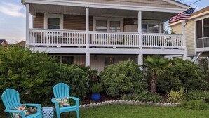 A two-story house with a balcony, a flag, and a garden with chairs and plants. - Pet friendly home w yard across from beach (Kure Beach)