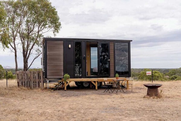 Situated in a rural setting, this compact home features a large sliding door and a cozy porch with a table and chairs.