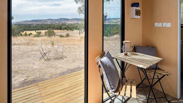 Una habitación con una mesa y sillas de madera, vista a un paisaje árido con árboles y colinas, y una mesita con una planta y una taza.
