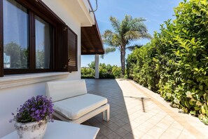 A patio with a white sofa and a potted plant, a tiled floor, and a view of a lush garden and palm tree. - Villetta Casetta Li Foggi (Gallipoli)