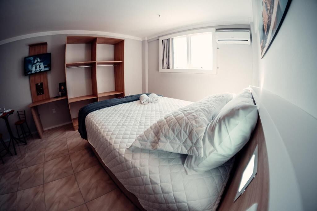 Une chambre à coucher comprenant un lit, un téléviseur fixé sur une étagère en bois, une fenêtre avec des rideaux et un appareil de climatisation.