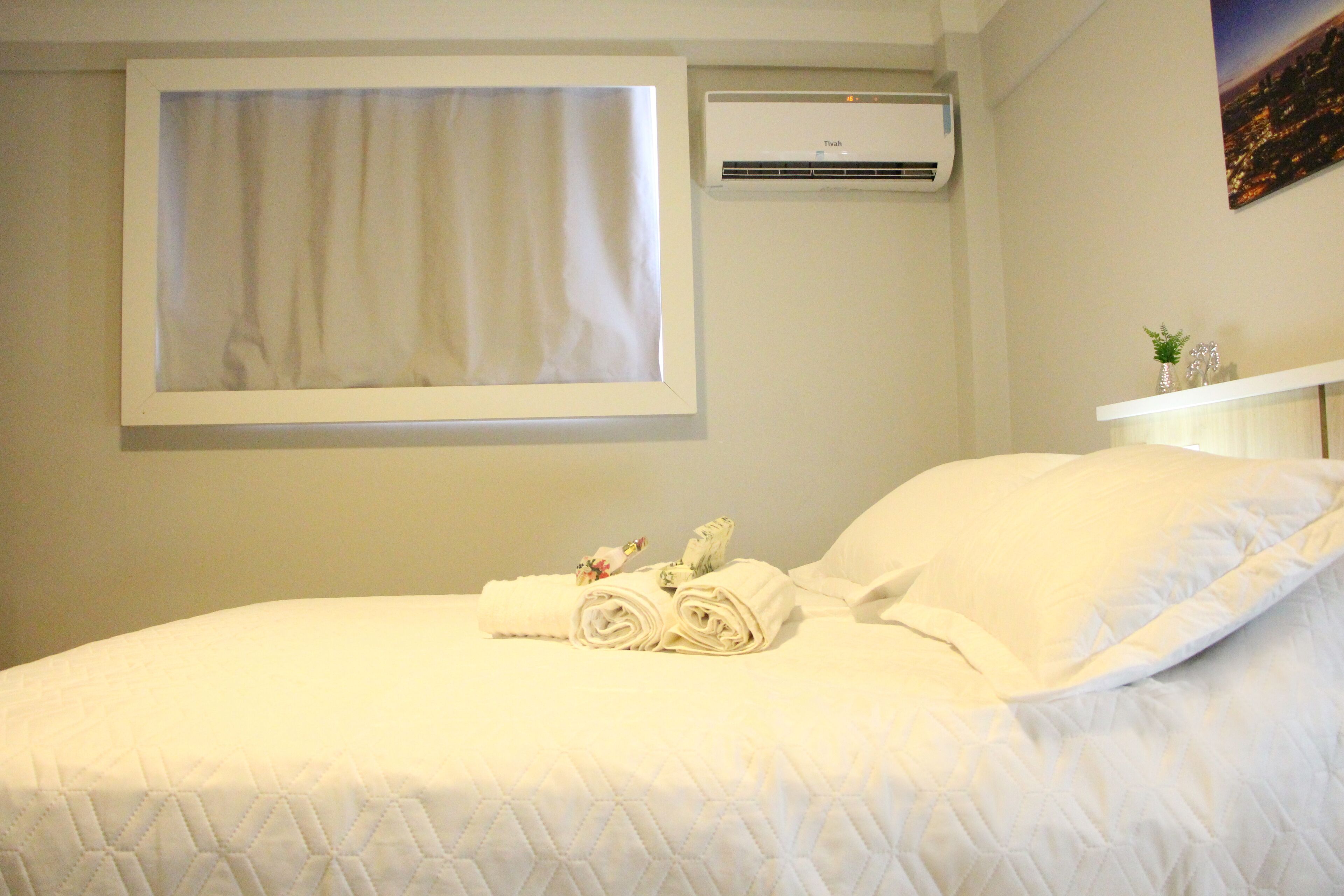 A neatly made bed with a quilted cover, a window with a white curtain, an air conditioning unit, and a small shelf with decorative items.