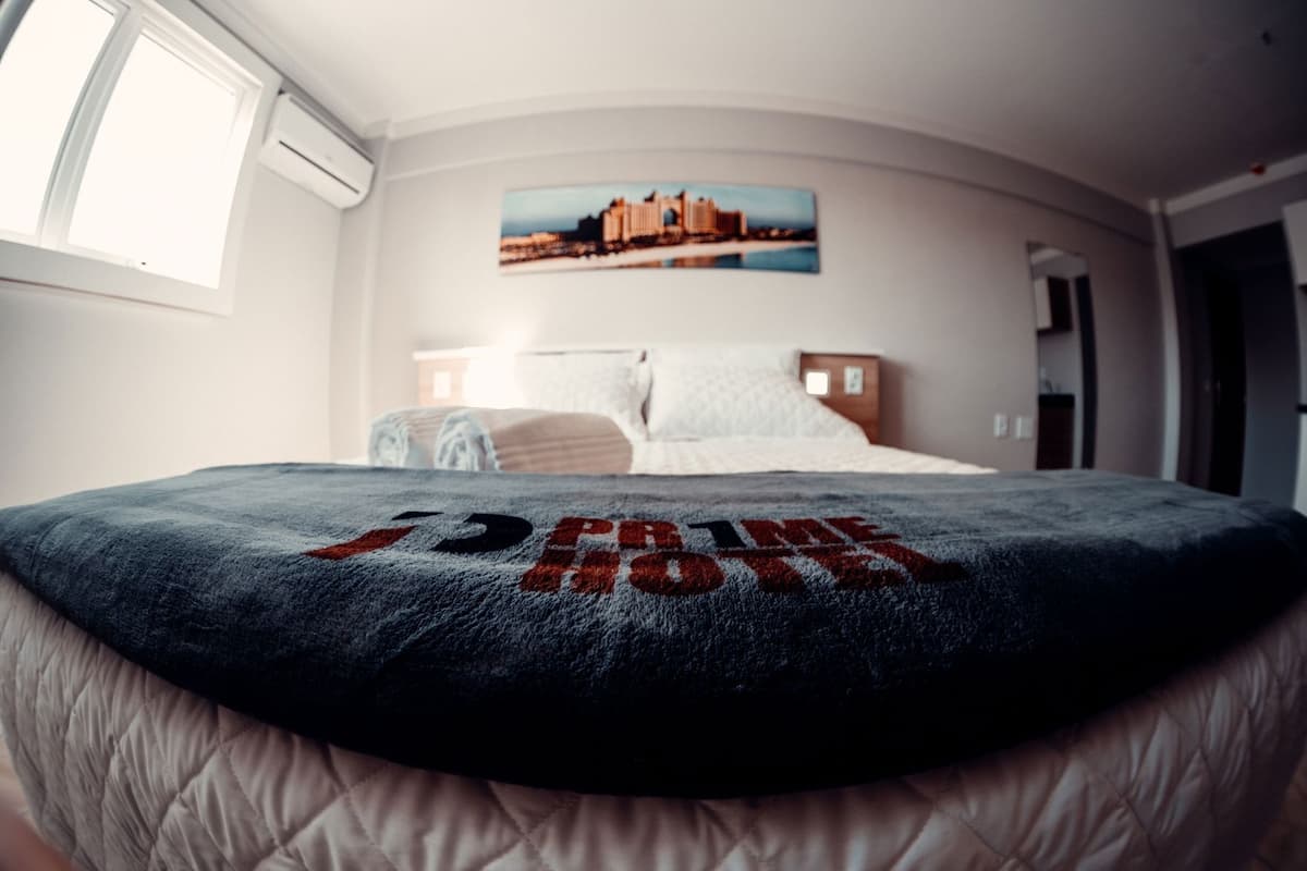 Un lit recouvert d’une couverture ornée du texte « The Ritz Hotel », une fenêtre et une image représentant un paysage urbain accrochée au mur.