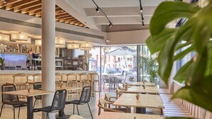 A modern cafe with wooden tables and chairs, a bar area, and large windows offering a view of the street. - Joe´s Gomila Boutique Hotel (Palma)