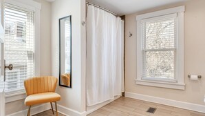 Una habitación con piso de color claro, cortina blanca, una silla y ventana con persianas.