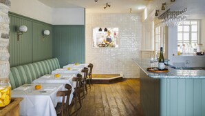 A restaurant with a wooden floor, green upholstered bench, white tablecloths, and a bar area with a wine bottle. - Cosy countryside retreat, edge of Frome near Bath. Pets welcome. (Mells, Frome)