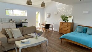 A modern living room with a sofa, coffee table, and a bed. There is a dining area with chairs and a table. A kitchen is visible in the background. - Cosy countryside retreat, edge of Frome near Bath. Pets welcome. (Mells, Frome)