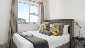 A neatly made bed with a grey headboard, white bedding, and decorative pillows. A window with curtains is visible, offering a view of the outdoors. - Two Bedroom Apartment | Pool | Netflix | Sea Views (Cape Town)
