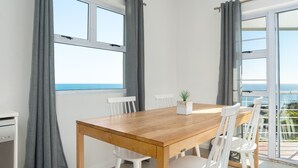 A dining area with a wooden table and white chairs, large windows with grey curtains, and a view of the sea. - Two Bedroom Apartment | Pool | Netflix | Sea Views (Cape Town)