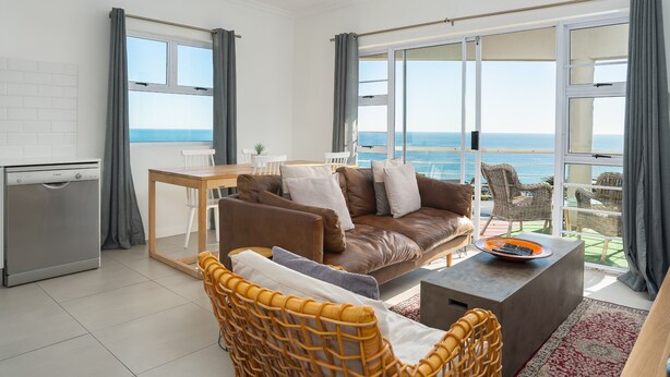 A modern living room with a sofa, a dining table, and a view of the ocean. - Two Bedroom Apartment | Pool | Netflix | Sea Views (Cape Town)