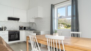 A modern kitchen with white cabinets, a wooden dining table, and white chairs. There is a window with a view of a mountainous landscape. - Two Bedroom Apartment | Pool | Netflix | Sea Views (Cape Town)