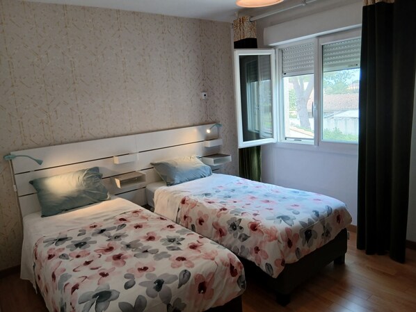 A bedroom with two beds, a window with blinds, a wall-mounted TV, and a bedside table with a lamp.