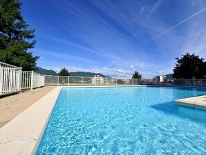 Piscina cercada por árvores e edifícios, com cerca branca, sob um céu claro.