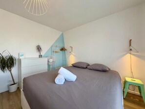 Quarto com cama, mesa de cabeceira, ventilador, planta pendente e um banquinho.