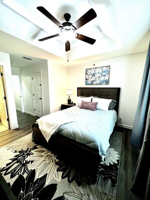 Quarto com cama, ventoinha no teto, mesa de cabeceira, abajur e tapete floral.