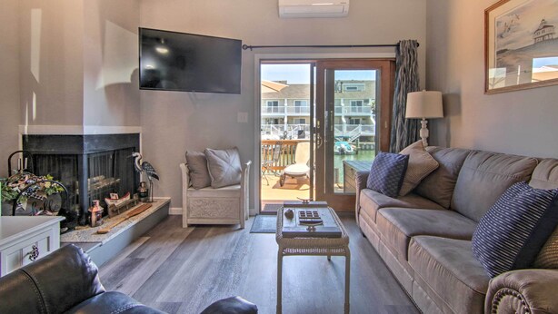 A living room with a fireplace, sofa, television, and a view of a balcony with outdoor furniture. - Pool Access, Near Beach! Waterfront Ocean City Gem (Ocean City)