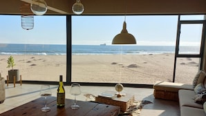 Habitació amb una finestra gran amb vista a una platja, una taula de fusta amb copes de vi i una ampolla, i una làmpada penjant.