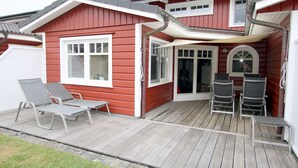 Ein rotes Haus mit einer Holzterrasse, zwei Liegestühlen und einem Tisch.