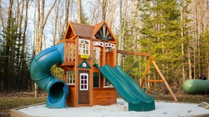 A wooden playhouse with a slide and swing set, surrounded by a sandy play area and trees. - Modern Mountain House in the Heart of Haliburton, Mins to Lake (Dysart et al)