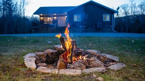 A campfire in a stone fire pit with a house in the background. - Modern Mountain House in the Heart of Haliburton, Mins to Lake (Dysart et al)