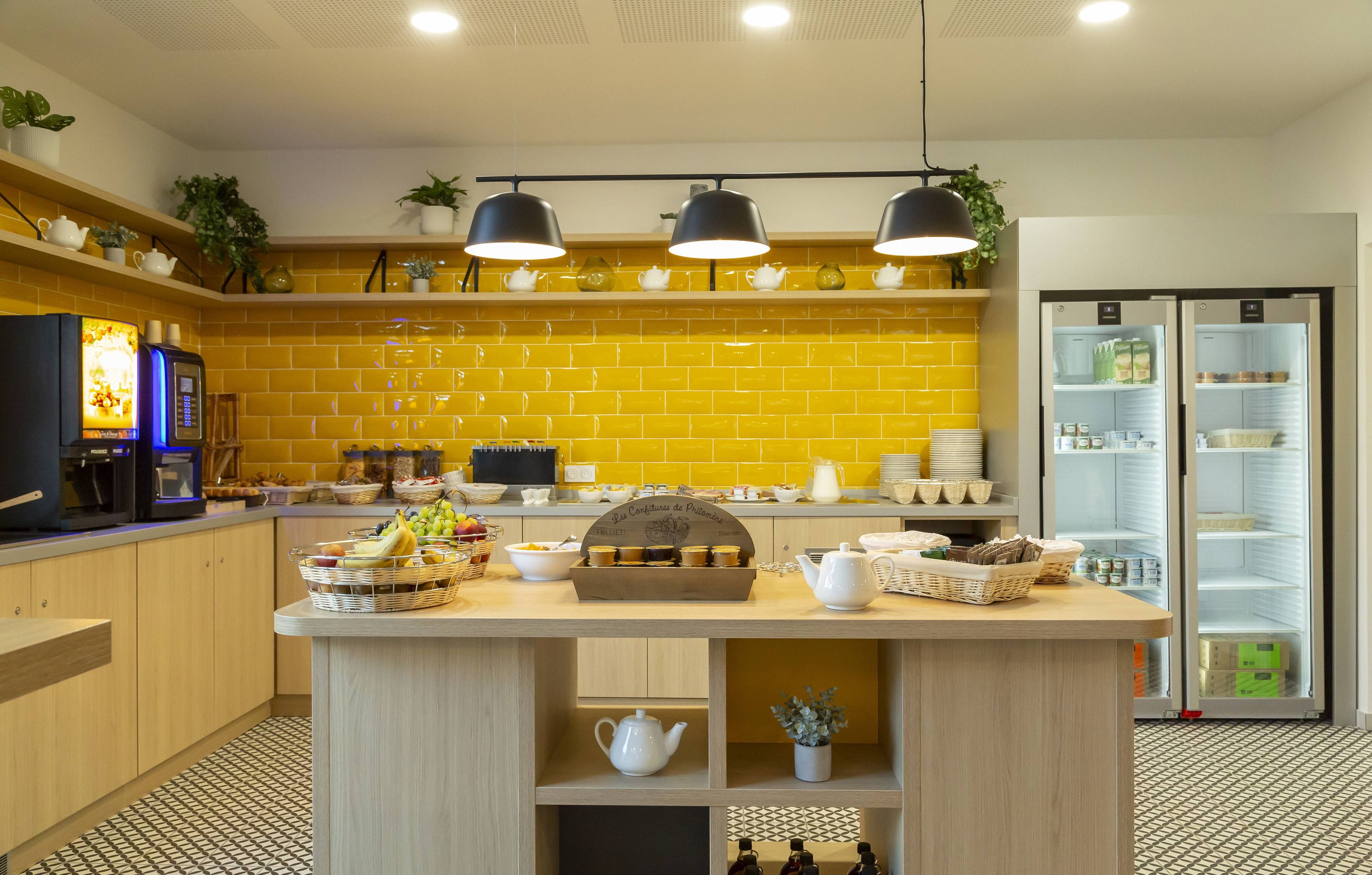 Eine moderne Küche mit gelben Fliesen, einer zentralen Kochinsel mit Teeservice, einer Kaffeemaschine und einem Kühlschrank.